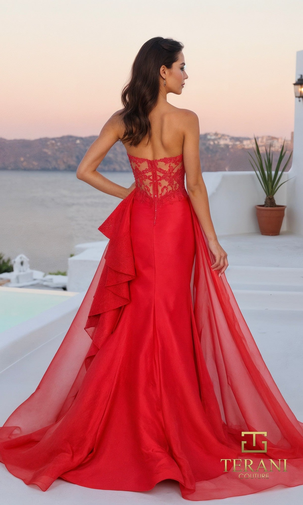 A woman stands outdoors facing away, wearing the Long Formal Dress: Terani 261P6261, a strapless Mikado gown with lace detail and flowing tulle overskirt. The scene features white architecture, a potted plant, and a waterfront view.